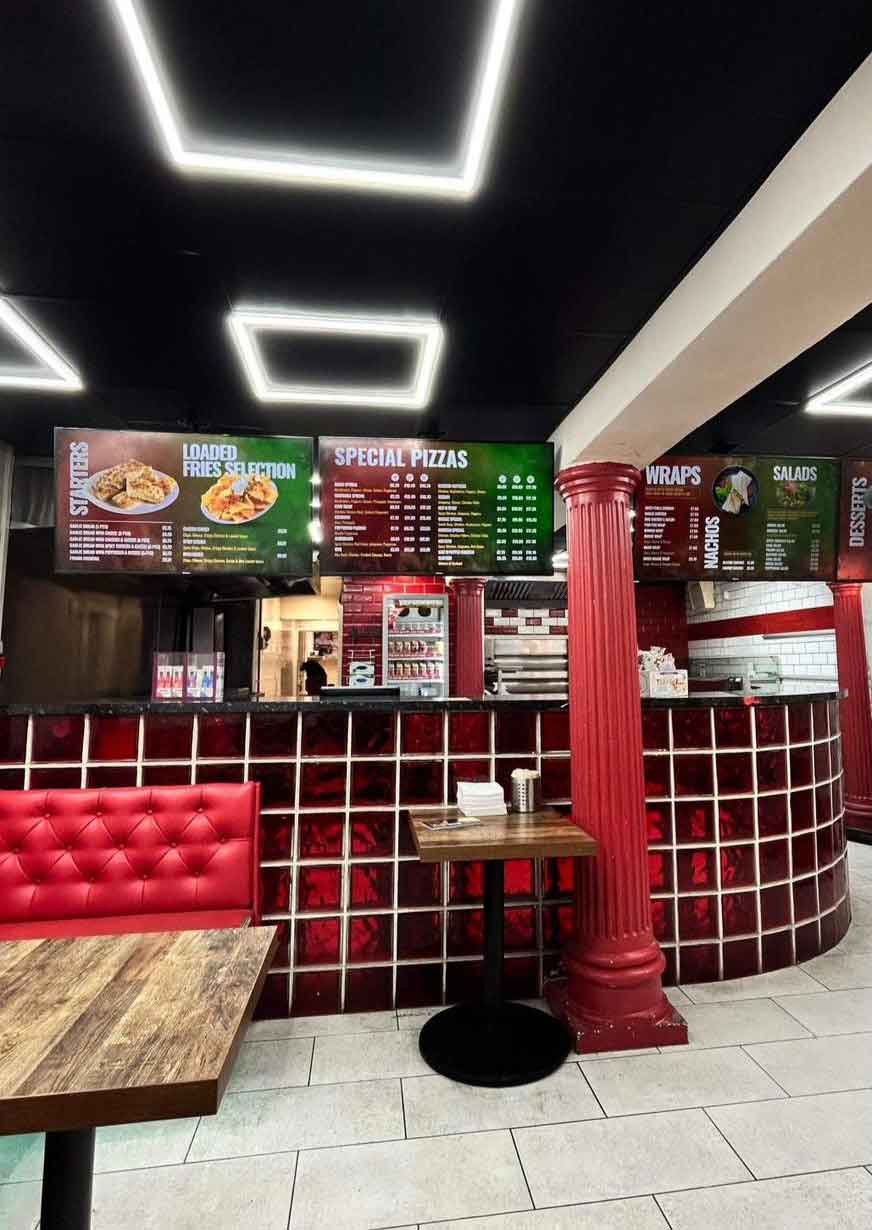 A Pizza shop counter with a red tiled base and a digital menu board overhead.
